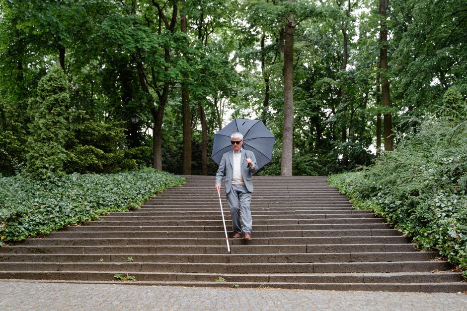 A man dressed in a grey suit, wearing sunglasses, walking down a set of outdoor stone stairs holding an umbrella in his right hand and a white cane in his left hand. The stairs are flanked by green leafy bushes on either side, with tall trees in the background, creating a shaded environment. The setting appears to be a park or landscaped area, and the man is navigating the descending steps carefully, likely as part of a home relocation or moving process coordinated by Man and Van Crofton Park, which specializes in removals and furniture transport services. The lighting indicates a possibly overcast day, emphasizing the use of umbrella and white cane for mobility assistance during a house move involving stairs and outdoor pathways.