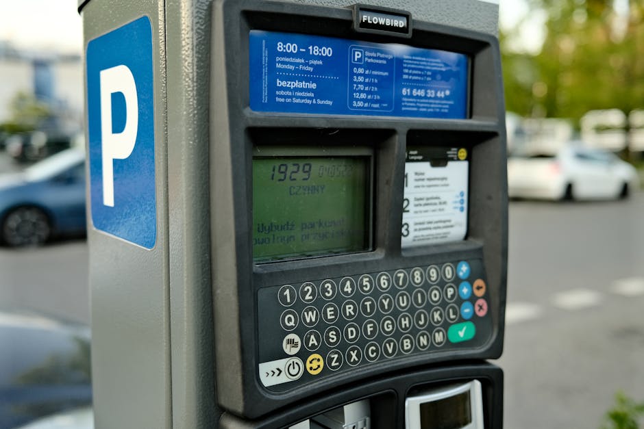 Close-up view of a parking meter situated on a paved street with several parked cars visible in the background. The meter displays a digital screen showing the time remaining and various instructions, with a keypad below it consisting of numbers, function keys, and symbols, including a smiley face and a checkmark. The parking meter is branded 'Flowbird' and is secured to the pavement, with part of a blue parking sign featuring a large white 'P' visible on its side. The surrounding environment includes a row of vehicles and some greenery, indicating a typical urban parking area. This image illustrates the process of parking payment, relevant in the context of house removals or moving services provided by Man and Van Crofton Park, especially when parking permits or payment at the loading/unloading zone are necessary during home relocation activities.