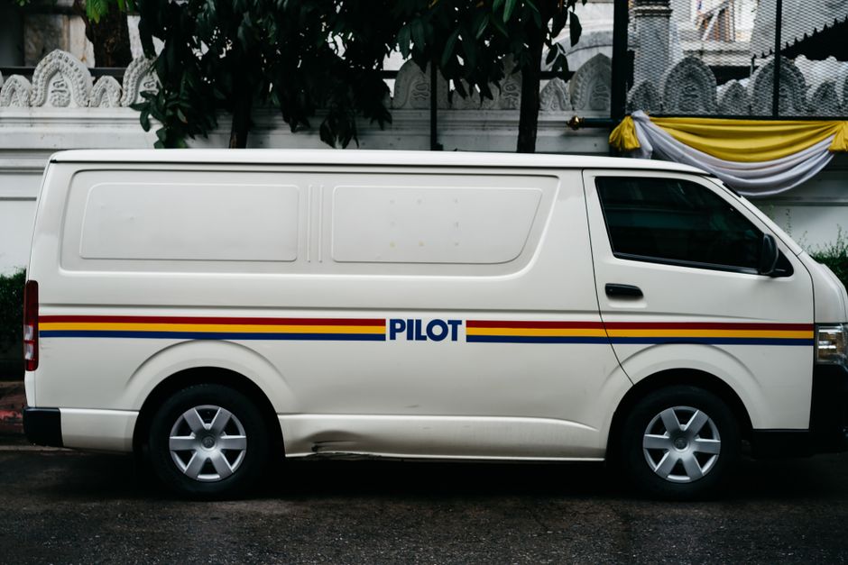 A yellow DHL delivery van is parked on a residential street, positioned partly on the cobblestone pavement and partly on the roadway, with its rear doors closed. Several other vehicles, including a bl