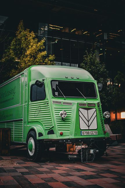 A vintage-style green moving van is parked on a brick-paved area outside a modern glass building at night, illuminated by surrounding artificial lighting. The van features a distinctive front grille, round headlights, and black side mirrors, with a license plate reading '6798 CC 15.' The vehicle is part of a home relocation or furniture transport service, associated with [COMPANY_NAME], and is positioned close to a loading zone. Visible inside the open back door are cardboard boxes, plastic-wrapped furniture, and packing materials, suggesting ongoing packing or loading activities. Nearby, a wooden pallet and various moving straps are seen, indicating the process of securing items for transport. The environment includes a small tree and urban surroundings, emphasizing the cityscape context of the move. This scene illustrates the logistics involved in house removals and the preparation for furniture transport during a moving or packing process.