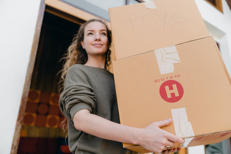 A young woman with long, curly hair and a neutral expression is standing indoors, holding two cardboard boxes stacked one on top of the other during a home relocation. One box has handwritten labels indicating it contains toys, while the other features printed packaging symbols and dimensions. She is wearing a casual, long-sleeved grey top with the sleeves rolled up. The background shows a part of a doorway and interior decor, with natural lighting illuminating her face and the boxes. The scene depicts the packing and loading process associated with professional removals services provided by Man and Van Crofton Park, highlighting packing materials like cardboard boxes and tape as part of furniture transport and house removals.