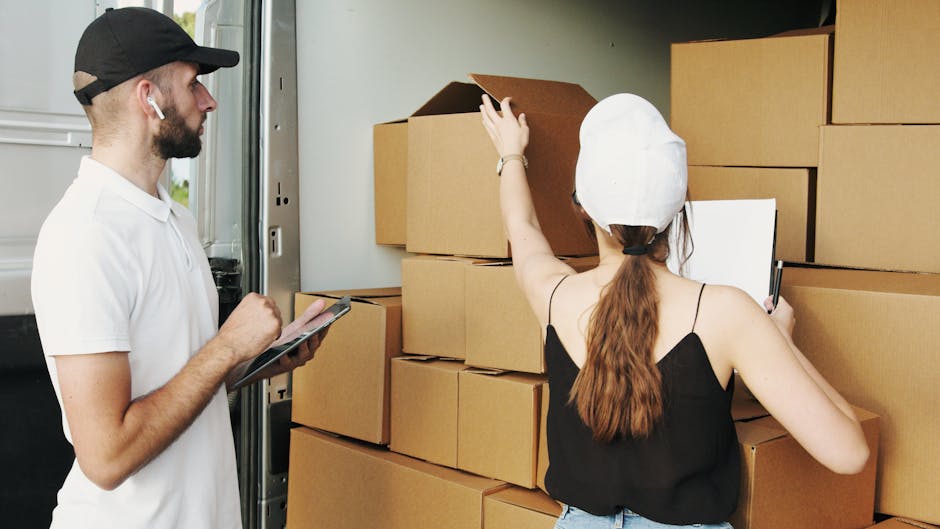 A man and woman engaged in a home relocation process next to an open moving van, with the woman standing inside the vehicle pointing to a stack of uniformly packed cardboard boxes on the ground. The m
