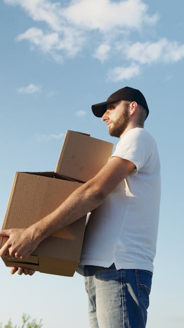 A man and woman engaged in a home relocation process next to an open moving van, with the woman standing inside the vehicle pointing to a stack of uniformly packed cardboard boxes on the ground. The m