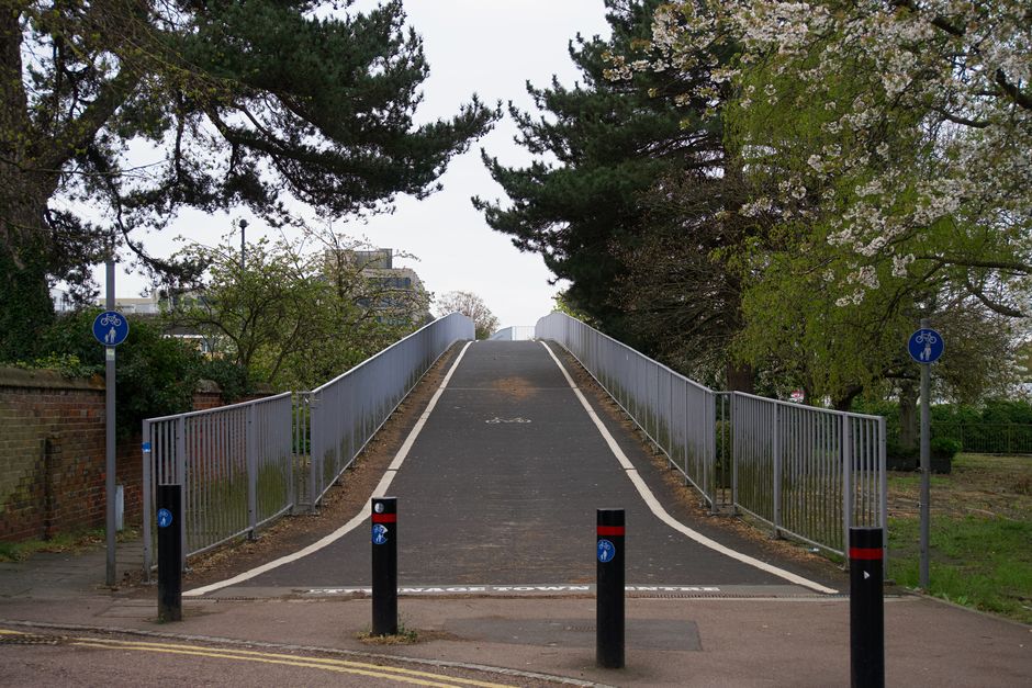 A straight asphalt pathway lined with leafless deciduous trees on both sides, stretching into the distance under a clear blue sky. To the left of the path, there is a large grassy area with minimal vegetation, and to the right, a fenced area with playground equipment and some structures. The trees cast long shadows across the path, indicating early morning or late afternoon sunlight. This outdoor scene, captured during a home relocation process, provides a clear view of the environment surrounding a residential area, with no visible furniture, boxes, or moving equipment. The image emphasizes an open, spacious setting typical of a street or parkway environment associated with house removals and relocation services provided by Man and Van Crofton Park, supporting the theme of transport logistics and loading processes along pavements involved in local moves and furniture transport.