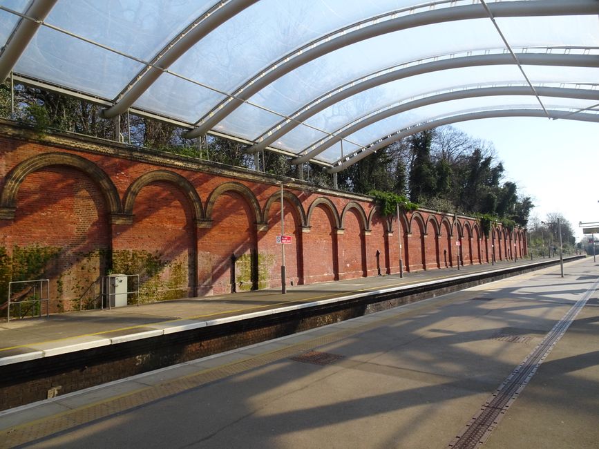 Empty train platform at Crofton Park Station SE4 with a curved translucent canopy overhead supported by metal beams. A long brick wall with arched brickwork runs along the back of the platform, partia