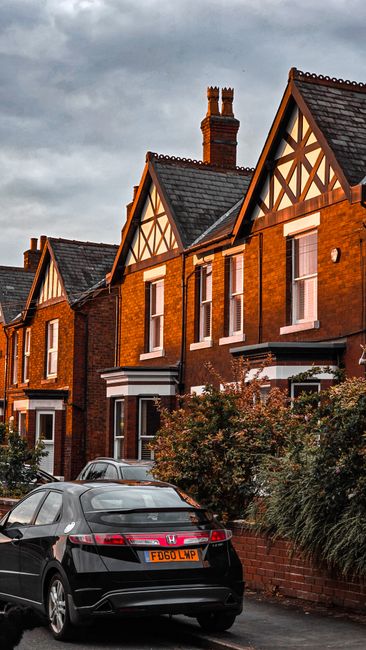 View of a residential street with a row of three attached brick houses under a clear blue sky, featuring pitched roofs with chimneys and multiple small windows. In the foreground, there is a parking a