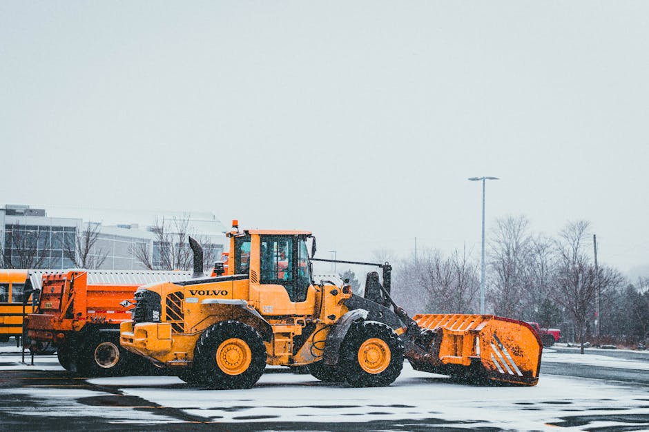 A bright yellow Volvo snow plough is stationary in an outdoor parking lot during a snowy day, with snow accumulating on the ground around it. The vehicle's large front blade, used for clearing snow, is tilted downward and partially covered in snow. The plough is positioned parallel to the paved surface, which is also lightly dusted with snow. In the background, there are leafless trees and modern buildings with glass windows, along with a tall street lamp and a few parked cars visible at a distance. The scene is overcast, with a grey sky suggesting cold weather conditions typical in winter. The snow plough's robust machinery and large tires are designed for heavy-duty snow removal, relevant to maintaining access during adverse weather when moving and transit logistics might be affected, similar to the considerations in house removals and home relocation services offered by Man and Van Crofton Park.