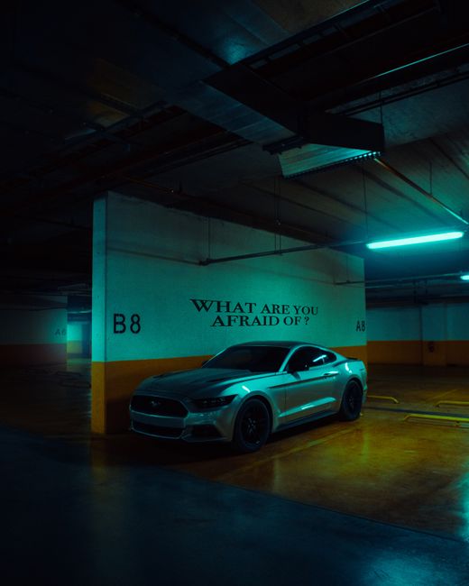 In an indoor underground parking garage with dim lighting illuminated by teal-colored fluorescent lights on the ceiling, a silver sports car is parked next to a concrete pillar marked with 'B8' and 'A8'. The pillar features a painted sign reading 'WHAT ARE YOU AFRAID OF?'. The car appears to be a modern coupe with tinted windows, positioned on a polished concrete floor that reflects the lighting. The garage's ceiling shows exposed pipes and ductwork, emphasizing an industrial environment. This scene may be part of a house relocation or moving process involving the transport of personal vehicles, as it relates to logistics surrounding household moves managed by companies like Man and Van Crofton Park.