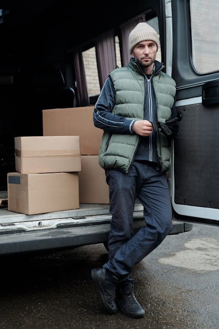A man dressed in a grey beanie, navy jacket with a green padded vest over it, and dark trousers, stands next to an open van doorway during a home relocation process. Behind him inside the van, there are several medium-sized cardboard boxes of varying heights, some sealed with packing tape, positioned on the vehicle's loading area. The man is holding a black glove in one hand, and appears to be taking a break from loading or unloading items. The van is grey and parked outdoors on a paved surface, with a building or house partially visible in the background. This scene captures the logistical aspects of furniture transport and packing during house removals, with visible objects and materials relevant to professional move logistics, as seen in the context of a house removal service at Crofton Park, as detailed on the website manandvancroftonpark.co.uk.