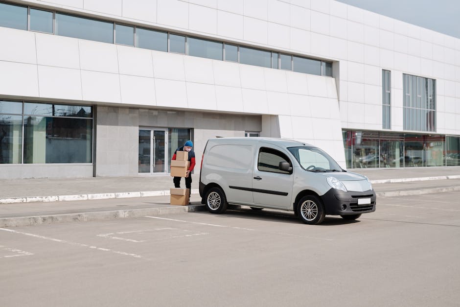 A person wearing a blue beanie and red jacket is standing outside a modern, white commercial building during daytime, loading four cardboard boxes onto a small silver cargo van parked in an empty parking lot. The boxes are stacked in a column, and the individual is in the process of placing them onto the vehicle's side. The van is positioned close to the curb, with the front facing slightly to the right, and the rear door open for easy access to load the boxes. The scene is well-lit with natural daylight, and the building features large glass windows and a sleek exterior design, typical of urban office or business premises. This image illustrates the logistics involved in home relocation or moving services, with a focus on packing and furniture transport, as provided by Man and Van Crofton Park, in relation to house removals and access tips for residential moves.