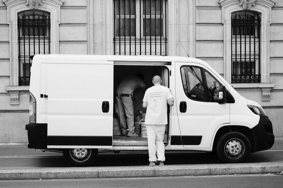 A man dressed in a grey beanie, navy jacket with a green padded vest over it, and dark trousers, stands next to an open van doorway during a home relocation process. Behind him inside the van, there are several medium-sized cardboard boxes of varying heights, some sealed with packing tape, positioned on the vehicle's loading area. The man is holding a black glove in one hand, and appears to be taking a break from loading or unloading items. The van is grey and parked outdoors on a paved surface, with a building or house partially visible in the background. This scene captures the logistical aspects of furniture transport and packing during house removals, with visible objects and materials relevant to professional move logistics, as seen in the context of a house removal service at Crofton Park, as detailed on the website manandvancroftonpark.co.uk.